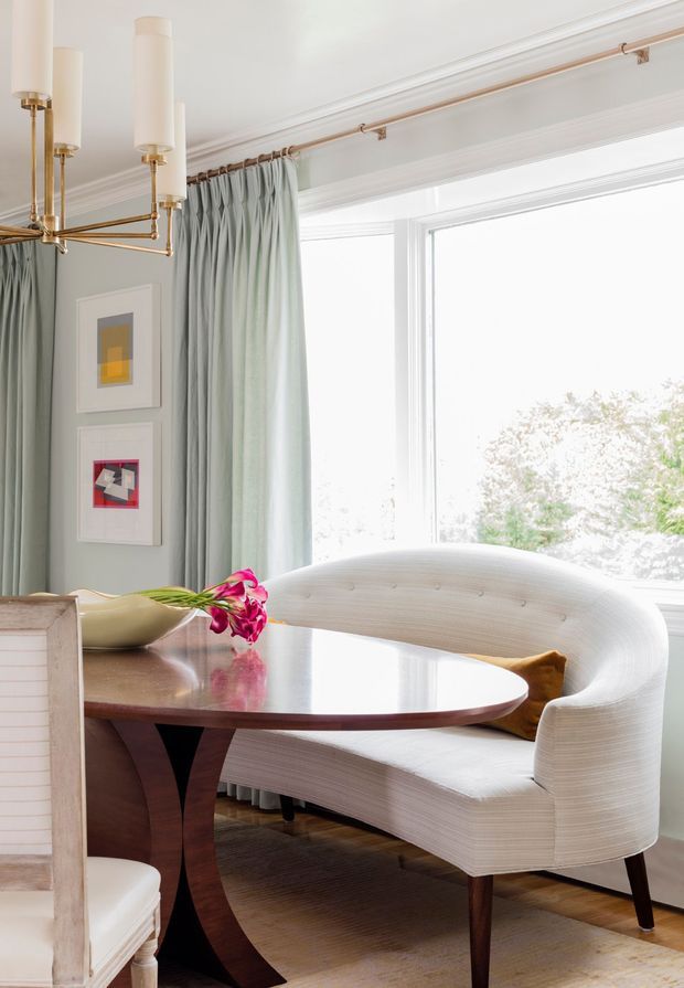 Bright, refined dining nook with a curved upholstered banquette, oval dark wood dining table, and partial light dining chairs. Pale blue-green floor-length curtains frame large windows, while a sculptural brass-and-cream pendant chandelier and framed artwork add decorative detail. The space has traditional moulded trim combined with contemporary furniture and a soft, airy palette.