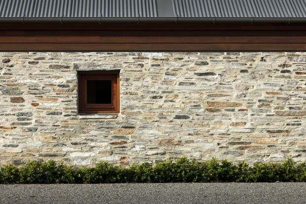 Close-up exterior view of a rustic stone masonry wall beneath a corrugated metal roof, with a dark timber-framed square window, horizontal timber detailing, and neatly trimmed hedging along the base.
