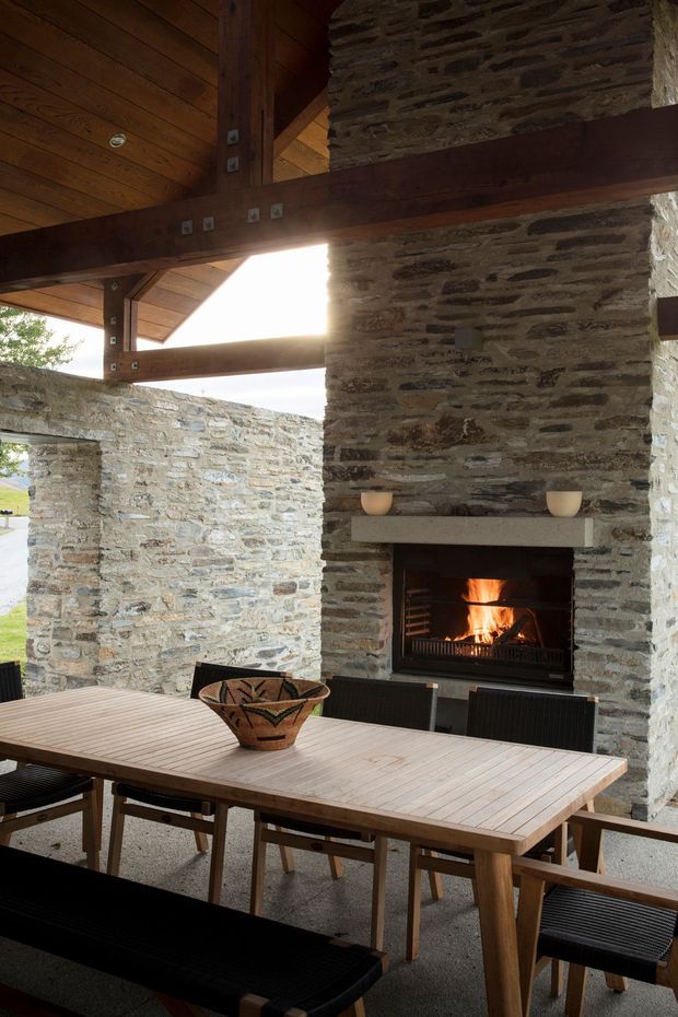 Covered outdoor dining area with a large timber dining table and benches, surrounded by rugged natural stone walls. A substantial open fireplace is built into the stonework, while exposed dark timber beams and a timber-lined ceiling create a warm, rustic architectural character.