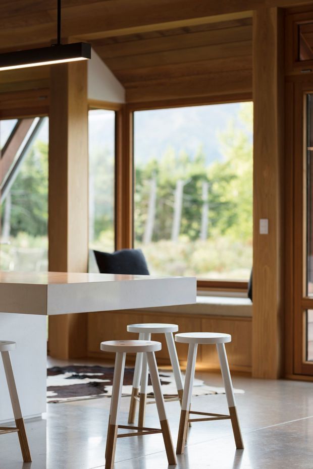 A warm, timber-lined open-plan kitchen or dining area with a white waterfall-edge island and three white-and-wood stools. Floor-to-ceiling timber-framed glazing provides views to greenery, while a black linear pendant hangs above the island. The room has a polished light concrete floor, exposed timber ceiling boards, integrated timber joinery, and a calm contemporary cabin-like character.