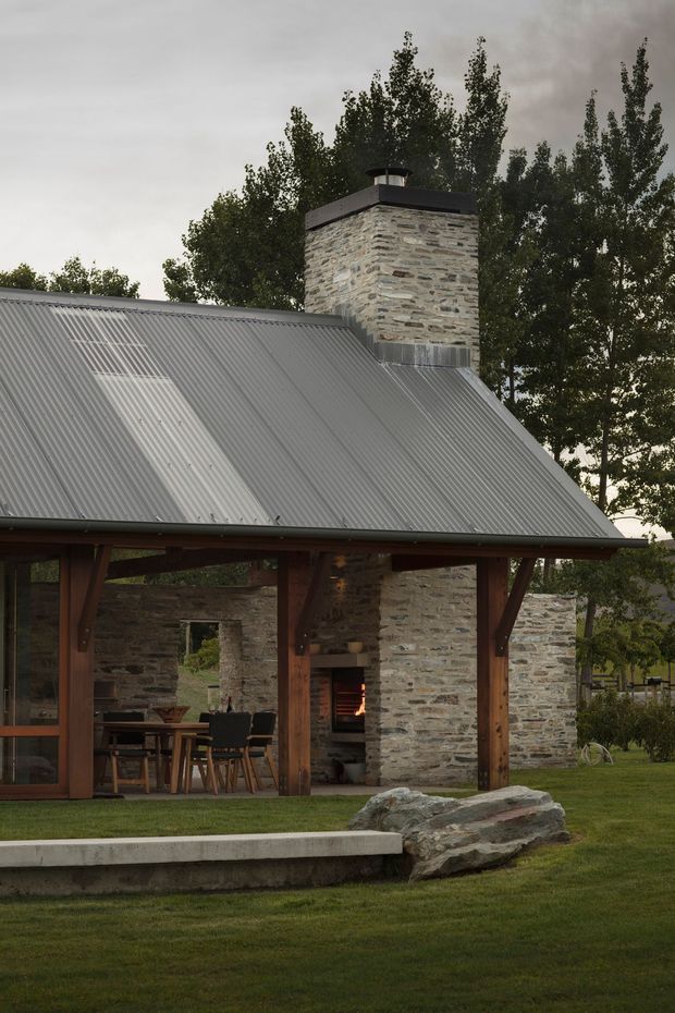 Exterior view of a rustic, single-storey pavilion-like house with a steep corrugated metal gable roof, exposed dark timber posts and beams, and a prominent rough natural-stone chimney. The covered outdoor area opens onto a lawn and contains a dining setting, while a fire burns in the built-in hearth. Mature trees and large natural boulders surround the building under an overcast sky.