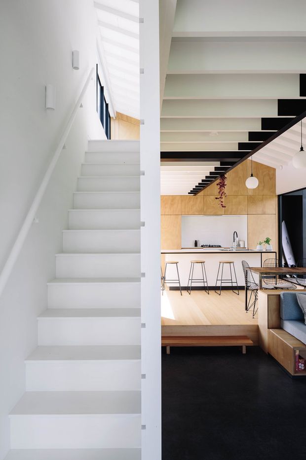 A bright contemporary open-plan home interior viewed beside a white staircase. The image shows a minimalist kitchen with pale timber cabinetry, white benchtops, bar seating, and a dining area, connected to a living room. Black structural beams and a black stair or mezzanine edge contrast with the predominantly white interior, while pendant lights and warm timber surfaces add visual interest.