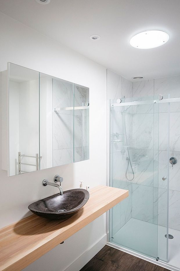 Bright contemporary bathroom featuring a frameless glass walk-in shower with large white marble-look wall tiles, a wall-mounted chrome tap, dark stone vessel basin, and a light timber floating vanity. Mirrored cabinetry spans the wall above the vanity, with recessed ceiling lights and a large oval flush-mounted light.