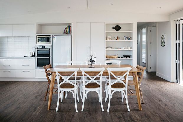 Bright, open-plan dining area adjoining a modern white kitchen. The room features a large timber dining table with mixed white cross-back and timber chairs, white built-in cabinetry, integrated appliances, open display shelving, dark timber-look flooring, and wide glazed doors opening to the exterior.