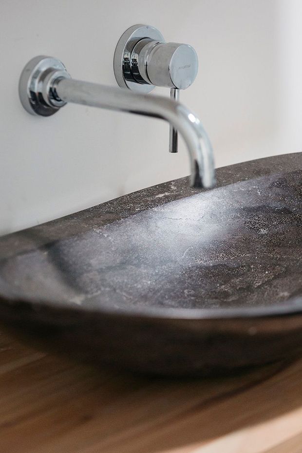 Close-up of a minimalist bathroom vanity featuring a wall-mounted chrome faucet above a dark, textured stone vessel basin. The basin sits on a warm timber vanity surface against a plain light wall.