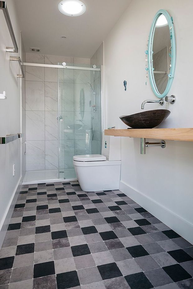Bright contemporary bathroom with a walk-in glass shower, wall-mounted toilet, vessel basin on a timber benchtop, oval aqua-framed mirror, pale marble-look wall tiles, and a bold checkerboard floor.