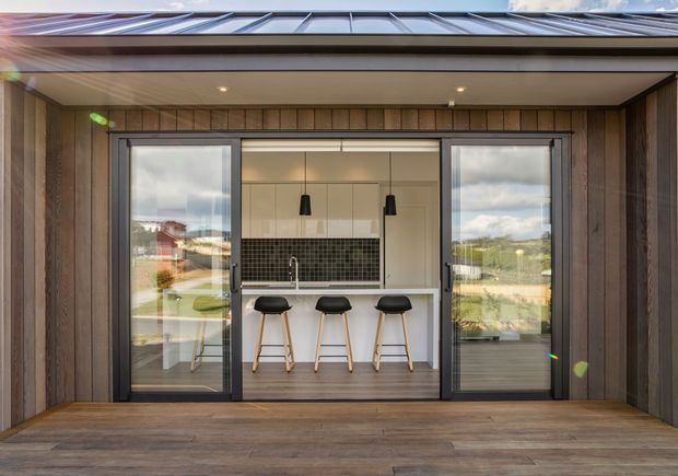 A contemporary covered outdoor deck or terrace looking into a compact kitchen through wide glazed sliding doors. The kitchen features a white built-in island with three black bar stools, dark square-tile splashback, pale cabinetry, and pendant lights. Dark vertical timber wall cladding, a timber deck, black-framed glazing, and a shallow roof overhang define the exterior. Reflections show the surrounding landscape and neighbouring buildings.