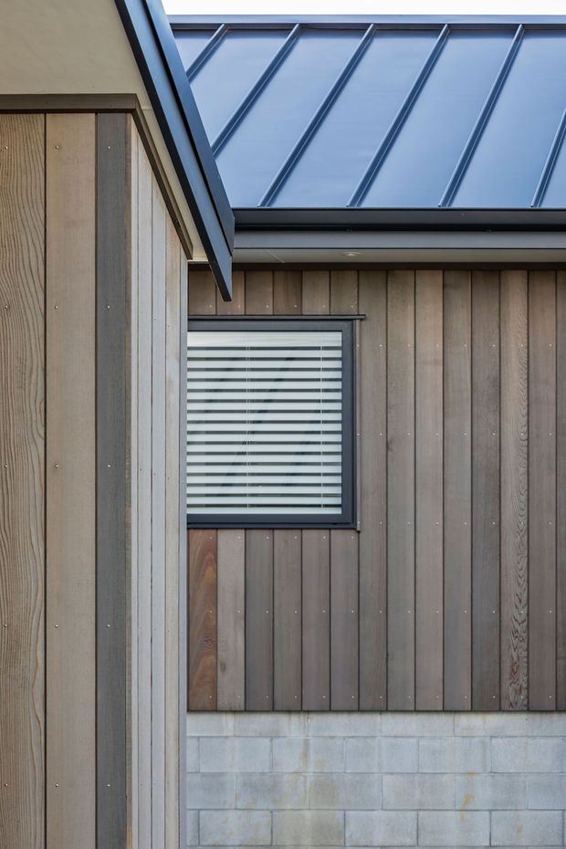 Close-up exterior architectural detail showing a modern building facade with vertical timber cladding, a grey concrete block base, dark metal window framing and horizontal blinds, and a standing-seam metal roof with pronounced ribs. The composition emphasizes the junction of roof planes and wall materials.