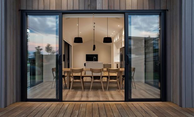 View from an exterior timber deck through wide black-framed glazed sliding doors into a dining area and adjacent open-plan kitchen. The interior features a timber dining table with matching chairs, pendant lights, pale cabinetry, timber flooring, and dark wood-burning fireplace or feature joinery. The exterior facade and deck are clad in vertical and horizontal timber boards.