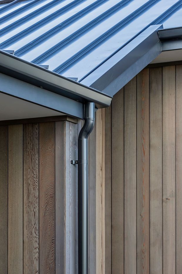 Close-up architectural detail of a building corner showing vertical timber wall cladding, dark metal gutters and downpipes, and a standing-seam metal pitched roof with carefully detailed eaves and junctions.