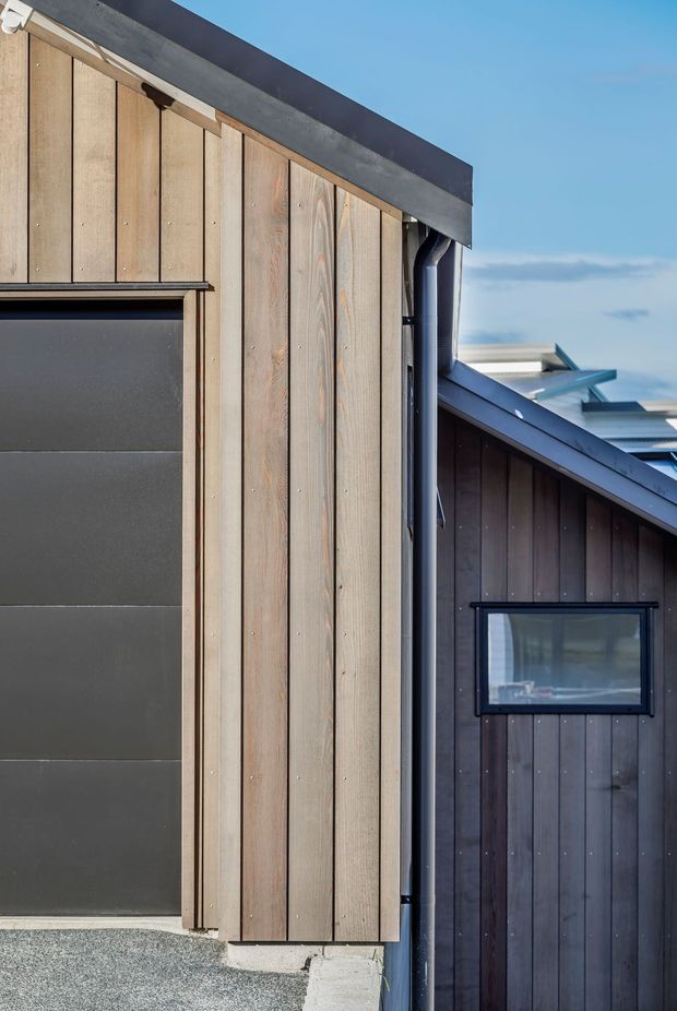 Close-up exterior view of a contemporary building with vertical timber cladding, dark metal roofing and gutters, and black-framed windows and doors. The composition highlights intersecting pitched roof forms and contrasting natural and dark-stained timber façades.