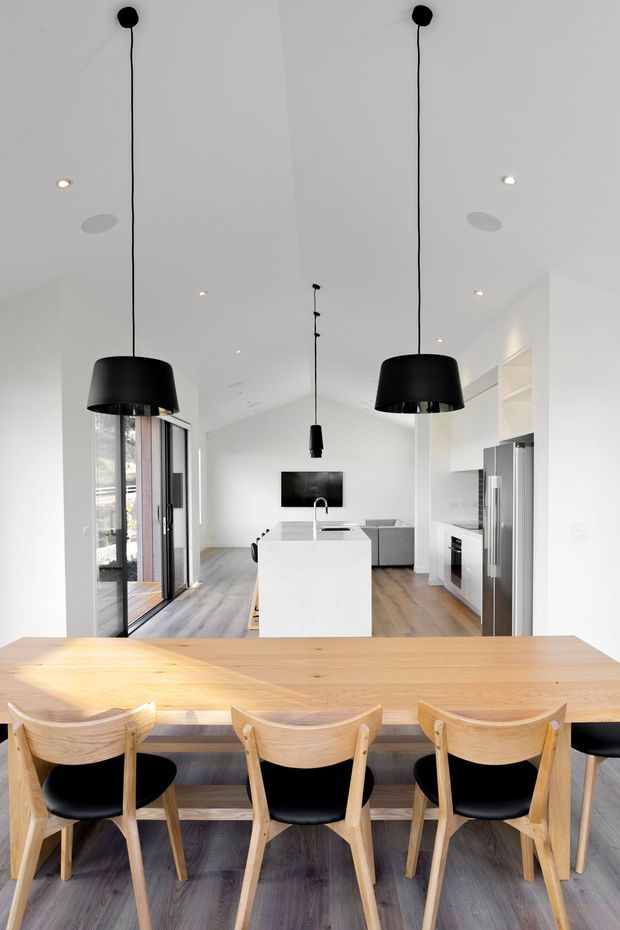 Bright open-plan contemporary interior viewed from the dining area toward a white kitchen and living space. A long light timber dining table with timber-and-black chairs dominates the foreground, while a white island, stainless steel appliances, pale timber flooring, large glazed doors, and oversized black pendant lights define the room. The space has a high pitched ceiling and a clean, minimalist aesthetic.