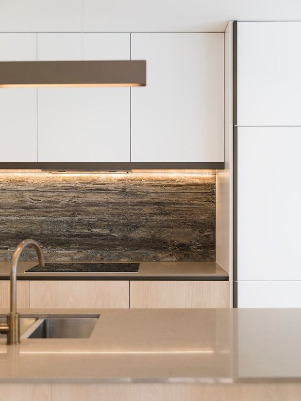 Close-up view of a refined contemporary kitchen with flat-panel white overhead cabinetry, pale timber lower cabinetry, a dark horizontally veined stone backsplash, integrated under-cabinet lighting, and a large warm neutral island with sink and bronze-toned tapware. The composition is minimal and highly detailed, with clean lines and concealed appliances.