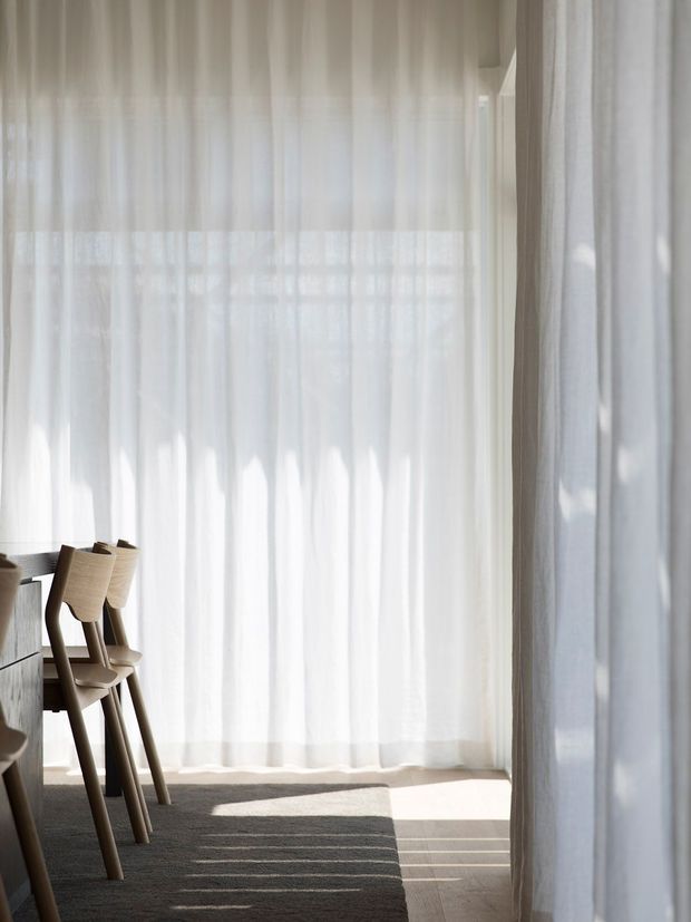 A softly lit dining area with floor-to-ceiling sheer white curtains, a light timber dining table and sculptural wooden chairs, and warm timber flooring. Strong sunlight filters through the curtains, casting elongated shadows across a neutral rug and floor. The restrained palette and uncluttered composition create a calm, airy interior.