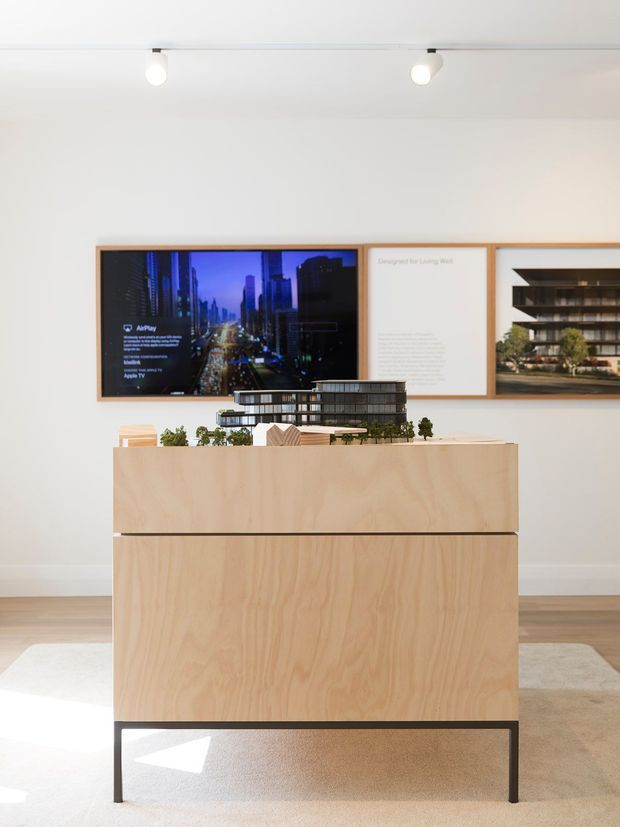Bright, minimalist showroom or presentation space featuring a large pale plywood display cabinet on black metal legs. Architectural models and small greenery sit on top, while framed architectural presentation boards and a digital display are mounted on the white wall behind. Timber flooring, a pale rug, and track lighting complete the clean exhibition-like setting.
