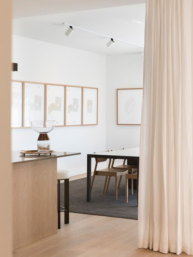 A bright, refined open-plan interior viewed through a doorway or curtain opening. In the foreground is a light timber kitchen island with a dark stone benchtop and a glass decorative bowl. Beyond it, a dining area contains a dark-framed rectangular table with timber chairs on a large charcoal rug. White walls display a series of pale timber-framed architectural drawings, while full-height cream curtains frame the right side. Track lighting and a clean, minimal material palette create an airy contemporary atmosphere.