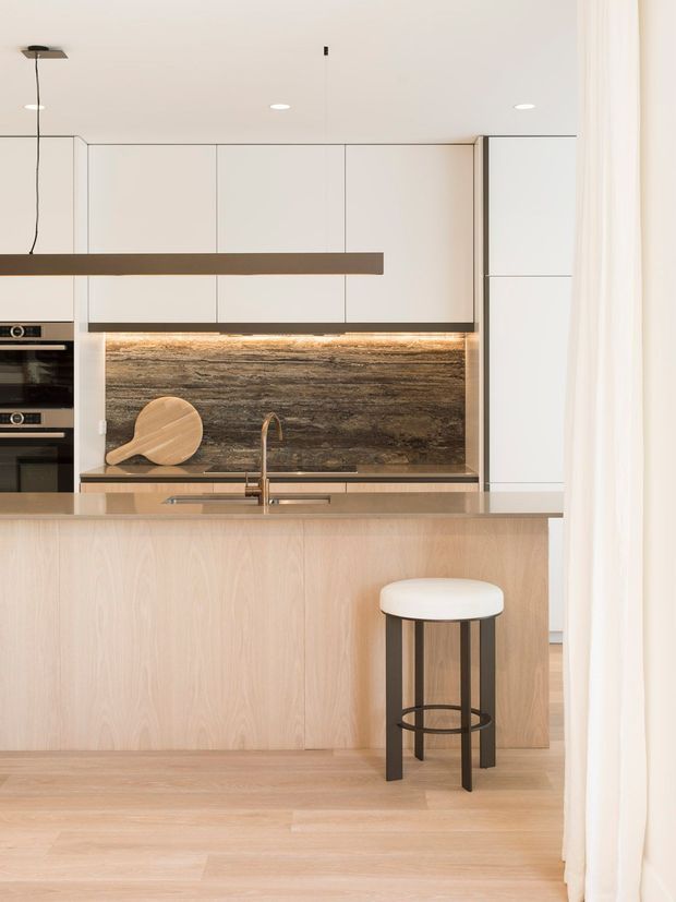 A bright, minimalist contemporary kitchen featuring a large pale timber island with an integrated sink, white handleless cabinetry, a dark textured stone splashback, built-in ovens, and a single bar stool. Soft sheer curtains and light timber flooring create a warm, refined atmosphere.