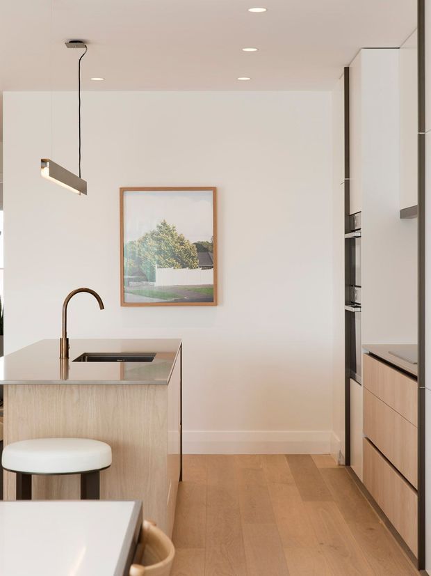 A bright, minimalist contemporary kitchen with a compact island featuring a pale timber veneer base, integrated dark sink and sculptural bronze-toned gooseneck tap. Full-height pale cabinetry lines the right side, with integrated appliances and warm timber drawer fronts. The space has light timber flooring, white walls and ceiling, recessed downlights, and a slim linear pendant. A framed landscape artwork adds a subtle natural accent.