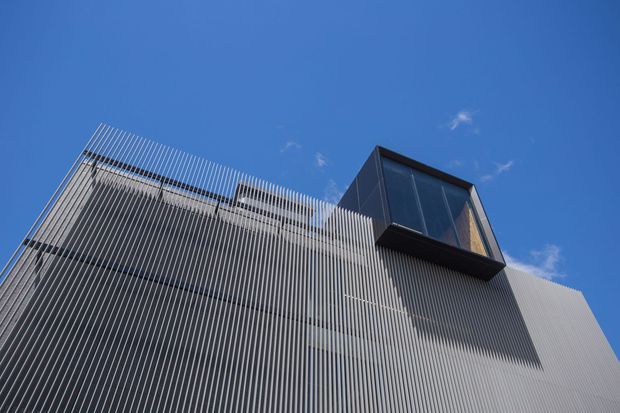 Low-angle exterior view of a contemporary building facade, dominated by closely spaced vertical fins or louvres in a light grey finish. A dark, projecting glazed upper volume contrasts with the screened facade beneath, set against a clear blue sky.