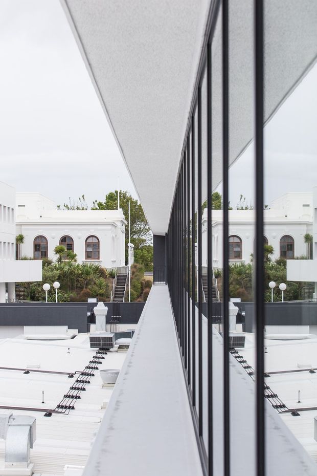 Architectural exterior view along a narrow elevated rooftop or terrace walkway beside a full-height black-framed glazed facade. The reflective glazing mirrors a white institutional or heritage-style building with arched windows, while the foreground shows a flat white roof, dark parapets, rooftop equipment, and landscaped planting beyond. The composition emphasizes strong linear perspective, symmetry, reflection, and the contrast between contemporary glazing and ornate white architecture.