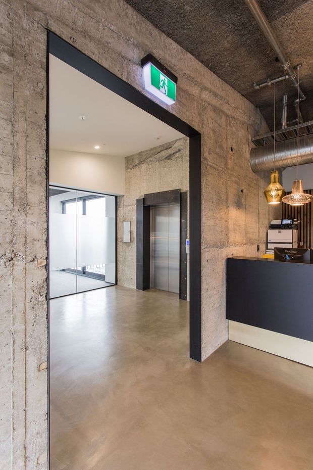 A commercial reception and lift lobby with an exposed industrial interior. The space features raw textured concrete walls, a polished concrete floor, exposed ductwork and services, a dark reception desk, pendant lights, and a stainless-steel elevator. A bright green emergency exit sign is mounted above the wide doorway.