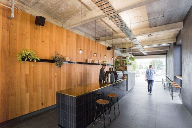 A modern cafe or hospitality interior with a long tiled service counter and bar seating, warm timber wall panelling, exposed concrete ceilings, suspended cable trays, pendant lights, industrial-style fixtures, and large glazing at the far end. Shelving with plants and small decor lines the timber wall, while additional stools and tables sit along the window side.