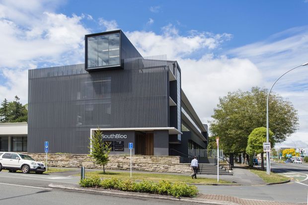 Street-level exterior view of a contemporary multi-storey commercial building with dark vertical ribbed cladding, extensive glazing, a projecting glass box, stone masonry at the base, and a recessed entrance marked SouthBloc. The building occupies a landscaped urban corner beside a roadway.