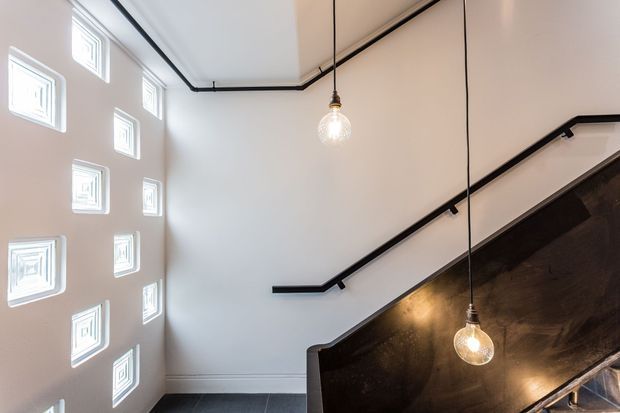 Interior view of a modern stairway with black metal handrails, dark timber or stained-wood stair detailing, suspended globe pendant lights, and a white wall punctuated by small square patterned windows. The composition is clean and architectural, with strong geometric lines and contrasting light and dark finishes.