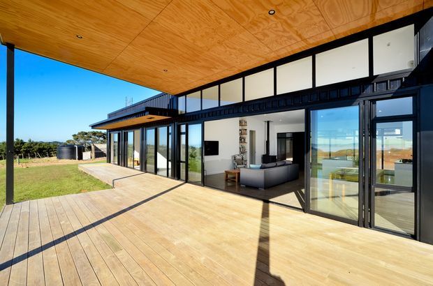 A contemporary single-storey home with extensive black-framed glazing opening onto a large timber deck. The covered outdoor area has a warm timber-lined ceiling with recessed downlights, while the interior living room is visible through the glass, featuring a grey sofa and white walls. The property sits in an open rural landscape with strong indoor-outdoor connection and expansive views.