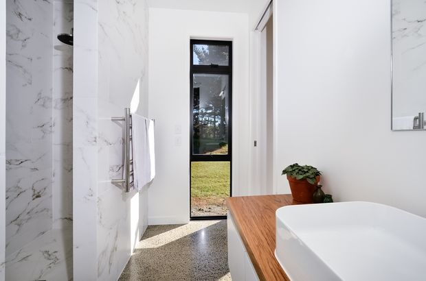 Bright contemporary bathroom featuring a freestanding white vessel bathtub on a warm timber vanity top, white marble-look wall tiles, polished metal towel rail, and a narrow black-framed glazed door opening to the outdoors. The terrazzo-style floor and abundant natural light create a clean, refined atmosphere.