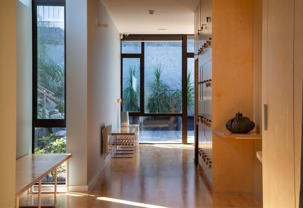 A bright, narrow residential hallway or entry passage with polished timber flooring, white walls, tall dark-framed glazing, and built-in timber cabinetry. The corridor leads toward a glazed rear opening with landscaped planting visible outside. A slim console table sits beside the window, while the cabinetry includes small display niches and hardware details. The space has a clean, warm, contemporary feel with strong natural light.