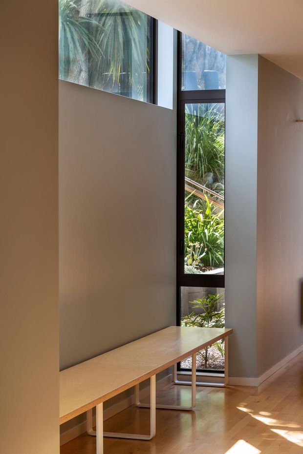 A narrow, light-filled interior passage or landing with a long built-in timber bench beneath tall, narrow black-framed windows. The glazing provides views onto dense greenery, while warm timber flooring and soft neutral walls create a restrained, contemporary atmosphere. Sunlight forms bright patches across the floor.