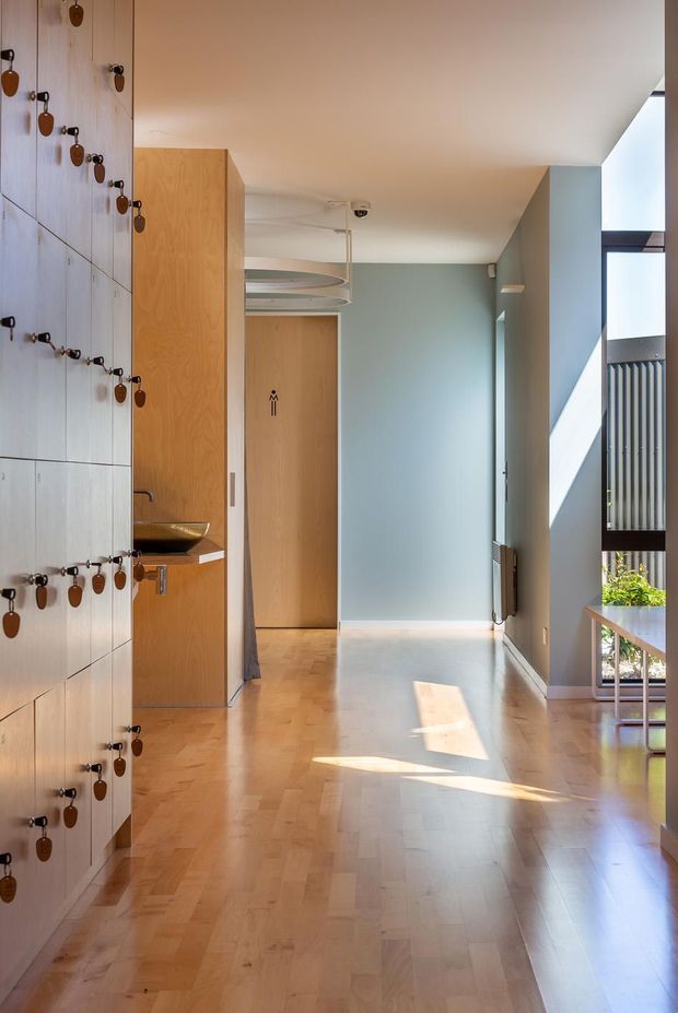 A bright, narrow residential hallway with pale timber parquet flooring, soft blue-grey walls, and extensive built-in timber locker cabinetry along the left wall. Each locker has a dark metal latch with a small leather or wood pull. A timber-lined vanity or basin niche sits midway along the cabinetry, while a timber door with a restroom symbol is visible at the end. Curved suspended ceiling elements and a large glazed opening on the right bring in strong sunlight, creating a clean, warm modernist interior.
