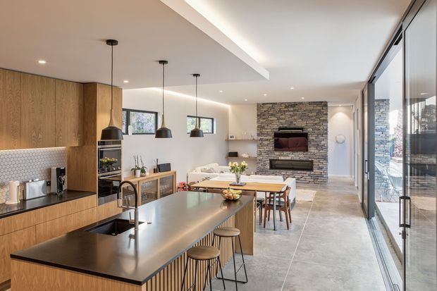 Open-plan contemporary kitchen, dining, and living area with extensive timber cabinetry, a large dark island benchtop, pendant lighting, and a stone-clad fireplace wall. Full-height glazed doors open to an outdoor area, creating a bright indoor-outdoor connection.