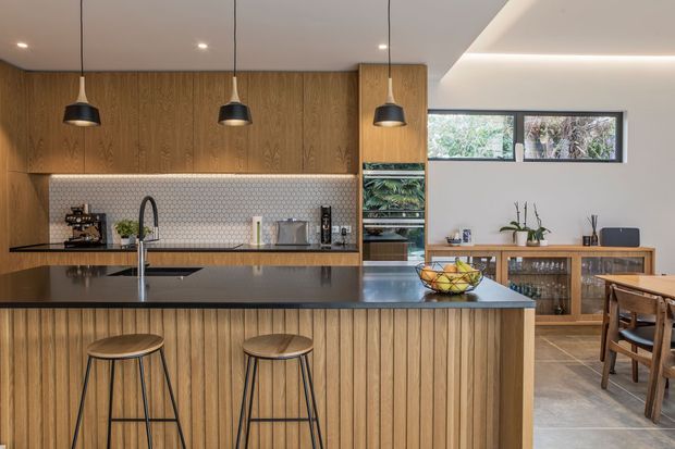 A warm contemporary open-plan kitchen and dining area. The kitchen features extensive vertical timber cabinetry and island panelling, a dark stone island benchtop with seating for two, a white hexagonal tiled splashback, integrated appliances, and three black pendant lights. A timber dining table and sideboard occupy the adjacent dining zone, with horizontal windows bringing in natural light.