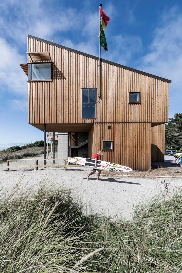 Exterior photograph of a compact, elevated timber-clad building set in a sandy coastal landscape. The asymmetrical multi-level form has a steeply sloping roofline, vertical natural timber boards, dark metal roof edging, small square windows, and a covered open area beneath the upper volume. Coastal grasses, a sandy path, a flagpole, and a person carrying a surfboard reinforce the beachside setting.