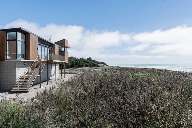 A modern multi-level coastal building set among low coastal vegetation beside a sandy shoreline and open ocean. The structure features dark timber cladding, grey masonry, extensive glazing, projecting balconies, and an external stairway, with a bright blue sky and distant headland beyond.