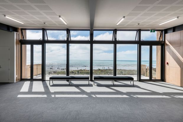 Bright contemporary communal interior with full-height black-framed glazing overlooking a sandy coastline and ocean. The room has grey carpet, pale timber wall panels, minimal black bench seating, linear lighting, and a perforated acoustic ceiling, with strong sunlight and window-frame shadows across the floor.