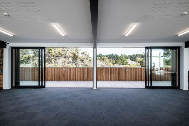 A spacious contemporary interior with a dark carpeted floor, white acoustic ceiling, exposed central structural beam, and large black-framed glazed sliding doors opening onto a timber deck. The room appears to be a flexible office or commercial space, with bright linear lighting and views to a fenced outdoor area and trees beyond.