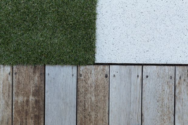 Close-up overhead view of a neatly arranged outdoor surface composition, with artificial-looking green turf meeting a speckled light concrete or stone section and a row of weathered vertical timber decking boards secured with visible dark fasteners. The image emphasizes contrasting textures and material junctions.