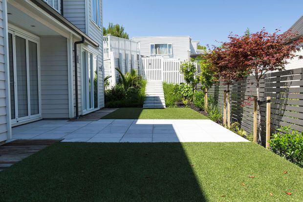 A bright, landscaped courtyard or narrow outdoor entertaining area between light-grey weatherboard buildings. The space features a tiled patio, lawn, planted garden beds, a slatted privacy fence, and a small set of steps leading to a raised deck or rear structure. White-framed glazed doors and horizontal weatherboard cladding are visible on the surrounding buildings.