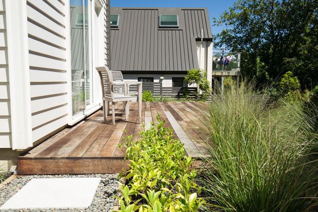 Sunny exterior view of a contemporary timber-clad home with a raised timber deck beside full-height glazing. Outdoor chairs and a small table sit on the deck, which transitions to a gravel path and landscaped planting with ornamental grasses and shrubs. A neighbouring dark vertical-clad structure is visible beyond the deck.