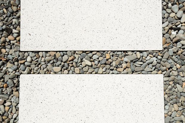 Top-down view of two large rectangular white concrete or stone slabs with a fine speckled finish, set among rounded grey, beige and brown decorative river pebbles. The image focuses on hardscape materials and geometric paving elements.
