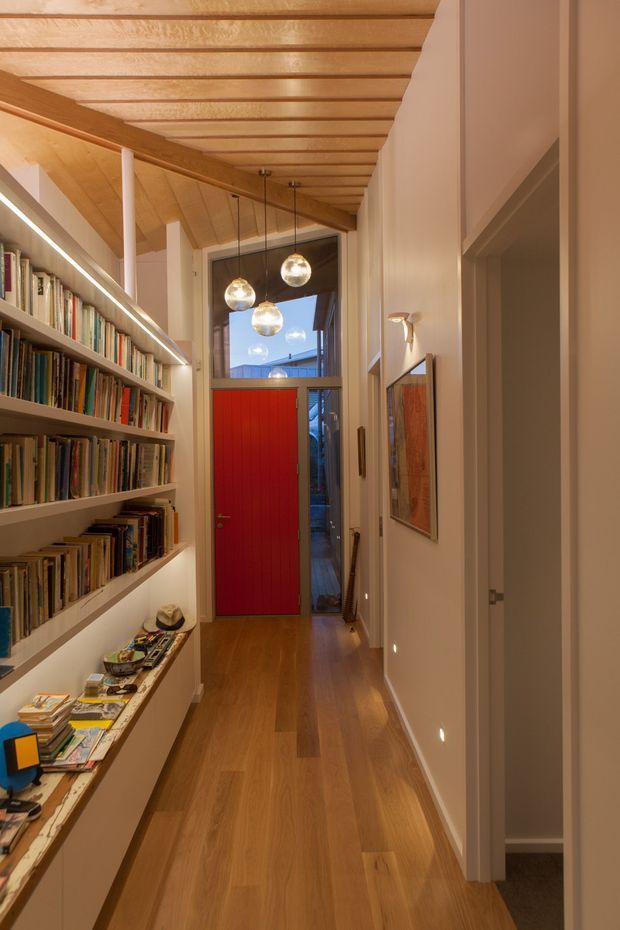 A narrow residential hallway lined with built-in open bookshelves and a low display bench on the left. Warm timber flooring and an exposed timber-lined ceiling create a continuous wood finish, while a bright red entry door anchors the far end. Pendant globe lights hang over the corridor, with framed artwork and recessed wall lights along the right-hand side. A partially open doorway reveals an adjacent carpeted room.