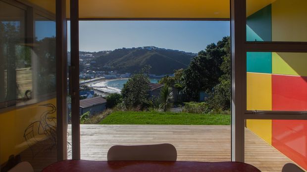 View from a bright interior dining area through full-height glazing and an open doorway to a timber deck, looking across a green coastal hillside and waterfront inlet. A colourful geometric panel in turquoise, yellow, and red frames the right side, while warm timber and dark-framed glazing define the space.