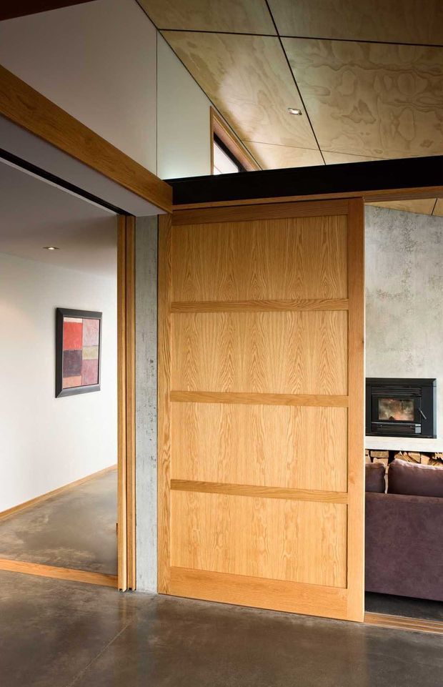 Interior view centred on a large natural timber pivot or sliding door with horizontal recessed panels. The door opens between a polished concrete-floored hallway and a living room containing a black fireplace and purple sofa. Exposed concrete wall sections, dark structural beams, timber-framed openings, and a plywood-lined ceiling create a warm, modern architectural interior.
