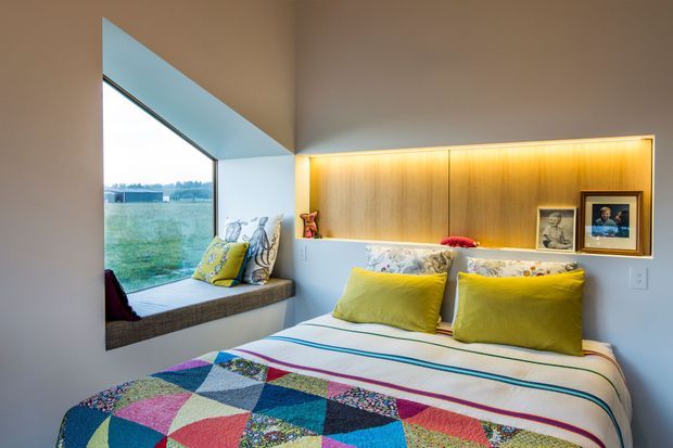 Bright, compact bedroom with a built-in window seat overlooking a green landscape. The bed is dressed in a colourful geometric patchwork quilt with striped bedding and lime-yellow cushions. A warm backlit timber wall niche behind the bed provides integrated display shelving for framed artwork and small decorative objects. The room has white walls, a large angled window, and a cheerful contemporary feel.