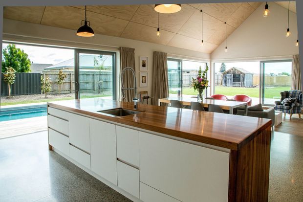 Bright open-plan kitchen centred on a large island with white handleless cabinetry and a substantial warm timber benchtop. The room has polished concrete flooring, a sloping timber-panelled ceiling, extensive floor-to-ceiling glazing, pendant lighting, and direct views to an outdoor pool and garden. A dining and living area is visible beyond the island.