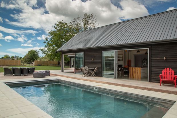Exterior view of a dark-clad, single-storey pavilion or house beside a rectangular swimming pool. The building has a steep corrugated metal gable roof, extensive sliding glass doors opening to an outdoor entertaining area, and a visible bar or kitchen zone inside. A paved pool terrace, outdoor dining setting, charcoal seating, and a bright red Adirondack chair create a relaxed contemporary outdoor space.