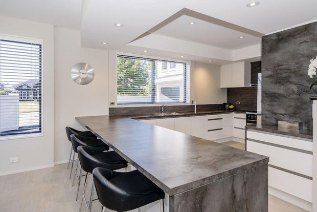Contemporary open-plan kitchen dominated by a large grey stone-look island with integrated seating for four. White flat-panel cabinetry, dark horizontal handles, dark tiled splashback, timber-toned benchtop surfaces, wide windows with horizontal blinds, recessed lighting, and a dramatic textured grey feature wall create a refined modern interior.